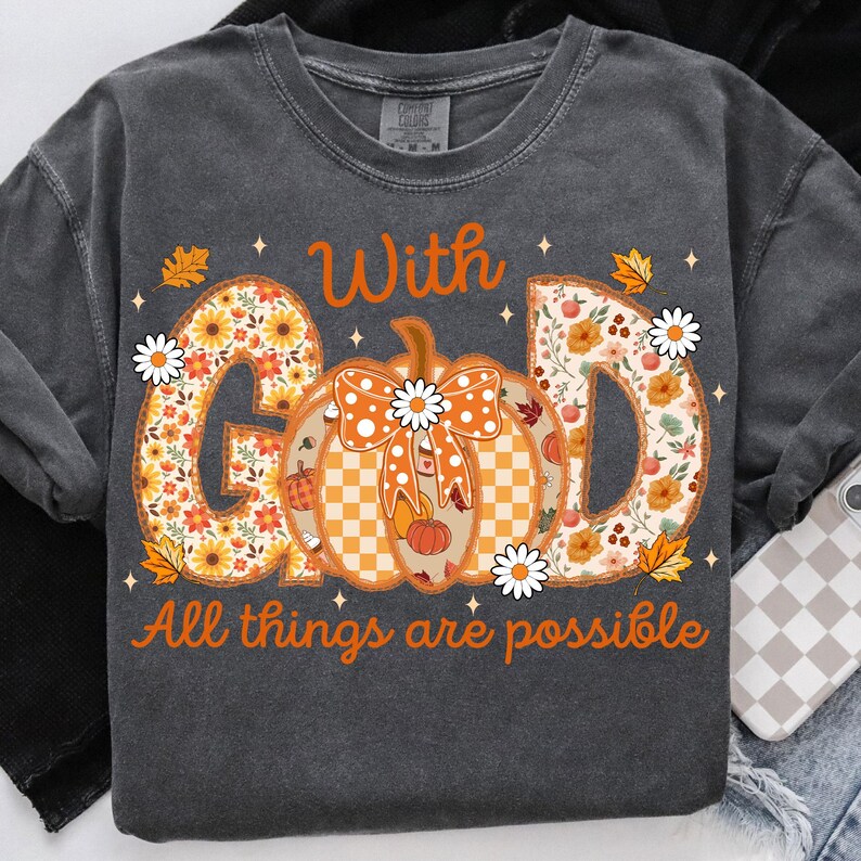 May include: Dark gray long-sleeve shirt with the words "With God All things are possible" in orange. The word "GOD" is filled with a floral pattern, and a pumpkin with a bow is in the center.