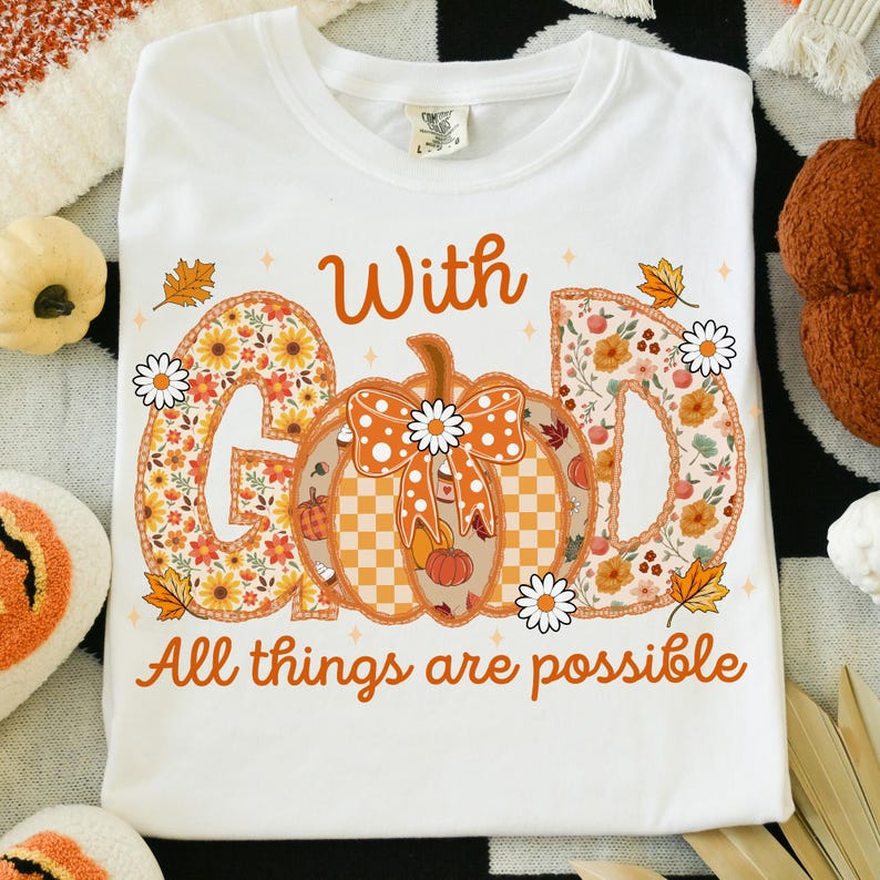 May include: White t-shirt featuring the words "With GOD All things are possible" in orange script. The word "GOD" is designed with floral patterns and a pumpkin in the center. Daisies and fall leaves decorate the design.