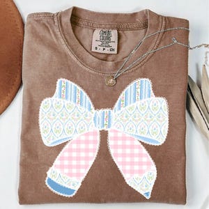 May include: A brown t-shirt featuring a large patchwork bow design in pink, blue, and white. The bow is made of different patterned fabrics, including gingham, floral, and striped patterns. The shirt has a crew neck and short sleeves. The text "COMFORT COLORS" is visible on the tag.
