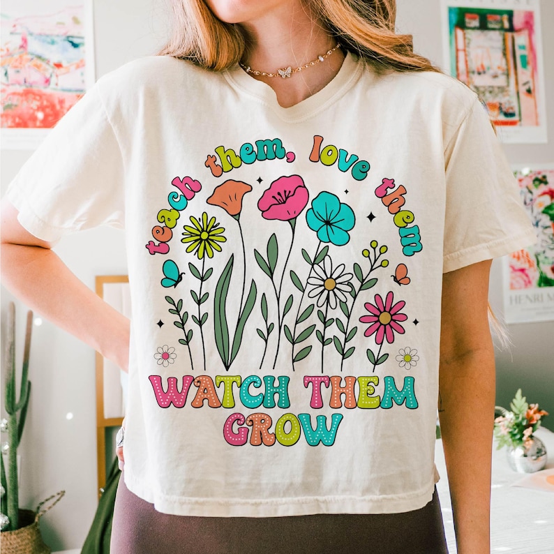 Puede incluir: Una camiseta blanca con un dise&ntilde;o gr&aacute;fico colorido que presenta flores y el texto "teach them, love them, watch them grow".