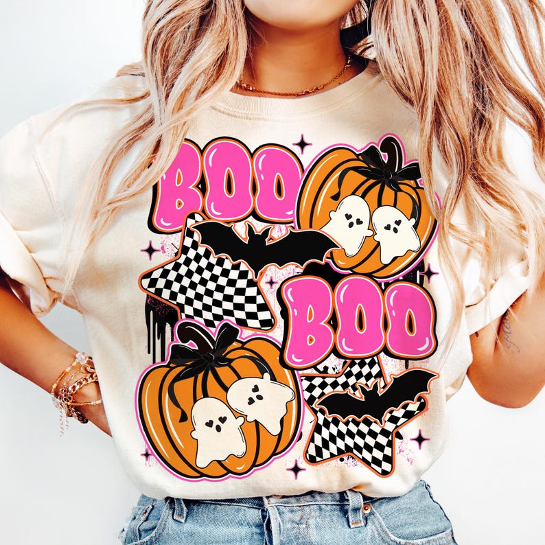 Puede incluir: Camiseta color crema con un dise&ntilde;o de Halloween. El dise&ntilde;o presenta la palabra "BOO" en letras rosas con burbujas, calabazas, fantasmas, murci&eacute;lagos y estrellas a cuadros. Las calabazas son naranjas.