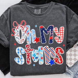 May include: Dark gray long-sleeve shirt with the phrase "Oh My Stars" in a patchwork design. The letters feature red, white, and blue patterns, including bows, stars, and gingham, with a few blue star accents.