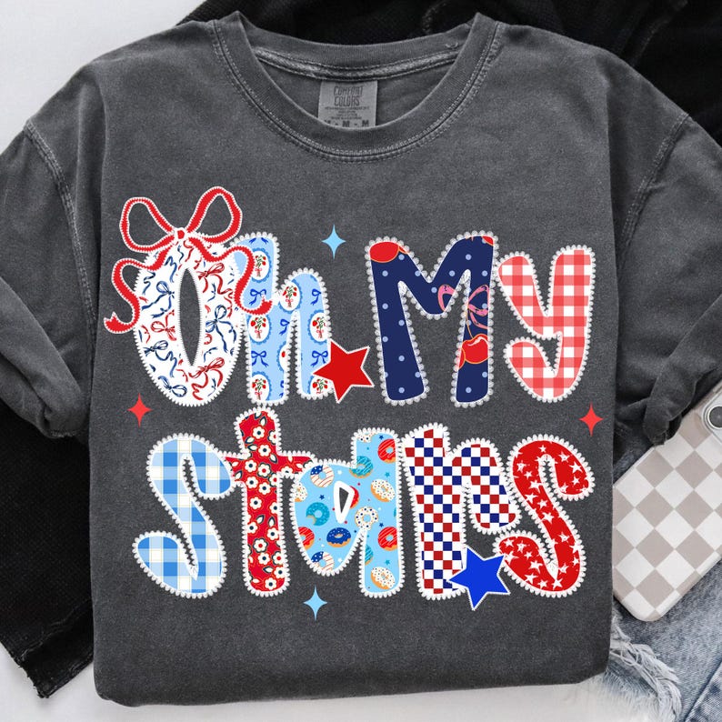 May include: Dark gray long-sleeve shirt with the phrase "Oh My Stars" in a patchwork design. The letters feature red, white, and blue patterns, including bows, stars, and gingham, with a few blue star accents.
