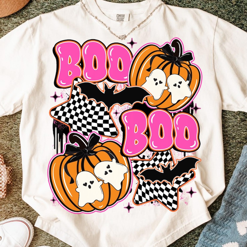 Puede incluir: Camiseta blanca con un dise&ntilde;o gr&aacute;fico de Halloween. El dise&ntilde;o presenta la palabra "BOO" en letras rosas, calabazas, fantasmas, murci&eacute;lagos y estrellas a cuadros. La camiseta tiene cuello redondo y mangas cortas.