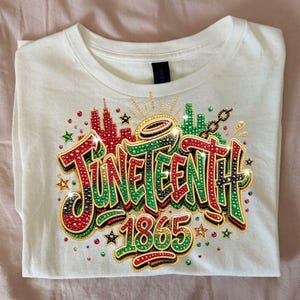 Pode incluir: Camiseta branca com um design Juneteenth colorido. O design inclui a palavra "Juneteenth" em vermelho, verde e preto, juntamente com o ano "1865". O design &eacute; embelezado com strass e estrelas.