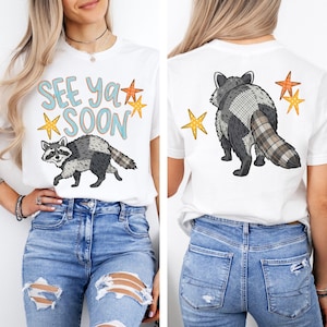 May include: White t-shirt featuring a raccoon graphic with the text "See Ya Soon" in blue and orange. The raccoon is depicted in gray and black with a plaid tail. The back of the shirt shows the raccoon's back and tail.