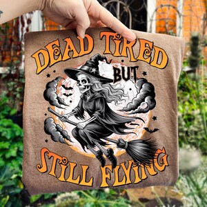 May include: A brown t-shirt featuring a Halloween-themed graphic. The design includes a skeleton witch riding a broom against a full moon, with the text "DEAD TIRED BUT STILL FLYING" in orange and yellow.