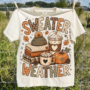 May include: A cream-colored t-shirt featuring the text "SWEATER WEATHER" in large, patterned letters. The graphic includes a stack of folded sweaters, a knit beanie, a coffee cup with a heart, and an orange pumpkin with small pinecones.