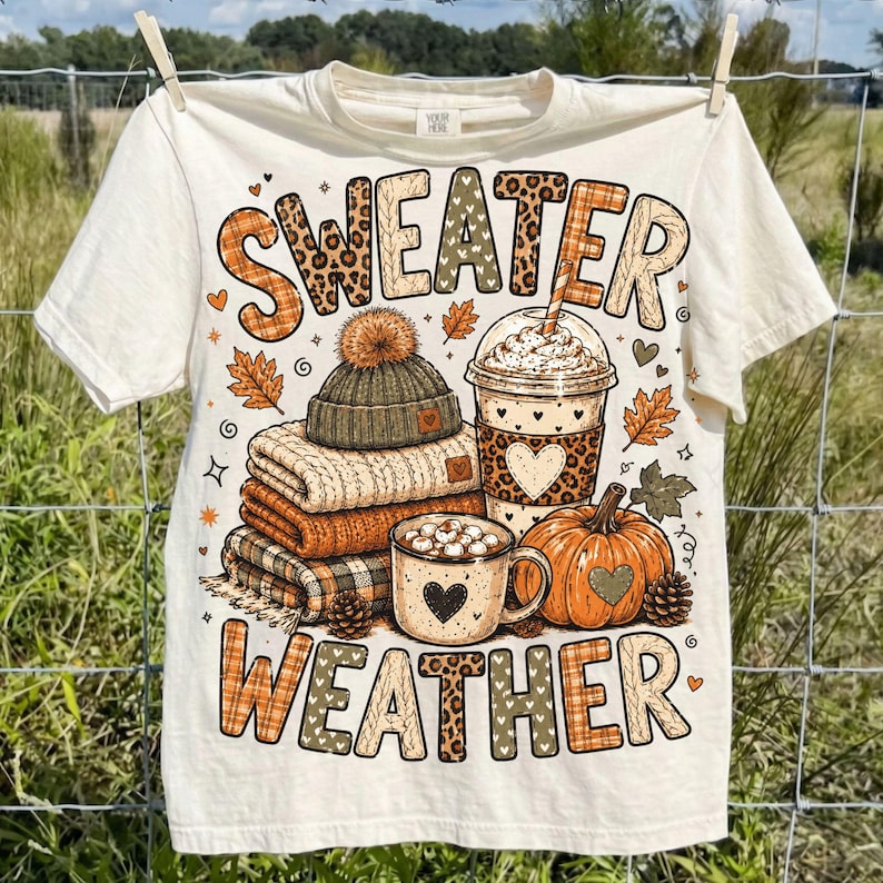 May include: A cream-colored t-shirt featuring the text "SWEATER WEATHER" in large, patterned letters. The graphic includes a stack of folded sweaters, a knit beanie, a coffee cup with a heart, and an orange pumpkin with small pinecones.