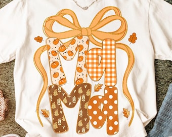 Op de afbeelding: Wit T-shirt met het woord "Mad" in oranje en bruine patronen, waaronder pompoenen, ruitjes en stippen. Een grote oranje strik en herfstbladeren versieren het shirt. Het shirt is van het merk Comfort Colors.