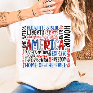 May include: White t-shirt with a patriotic design. The design features the word "AMERICA" in red and blue, surrounded by phrases like "Liberty," "Freedom," and "Home of the Free." The shirt is perfect for celebrating Independence Day.