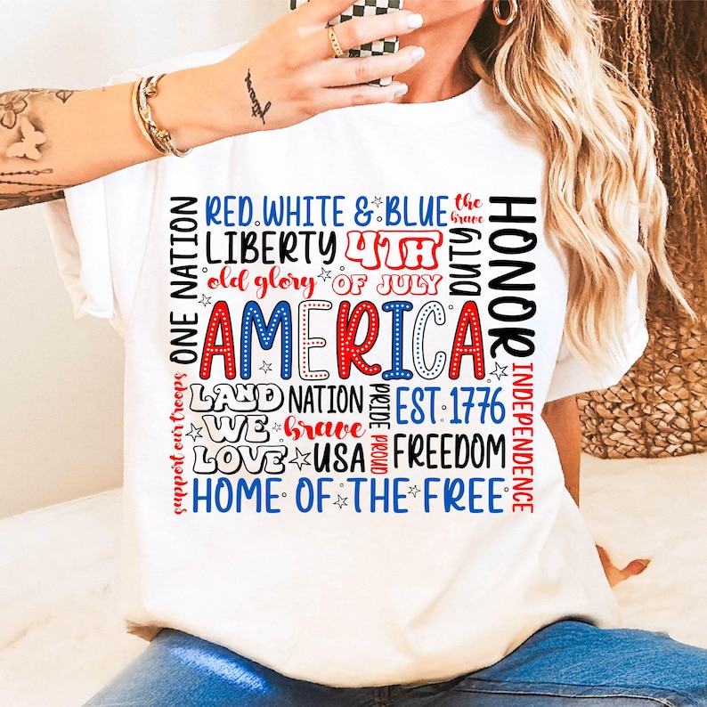 May include: White t-shirt with a patriotic design. The design features the word "AMERICA" in red and blue, surrounded by phrases like "Liberty," "Freedom," and "Home of the Free." The shirt is perfect for celebrating Independence Day.