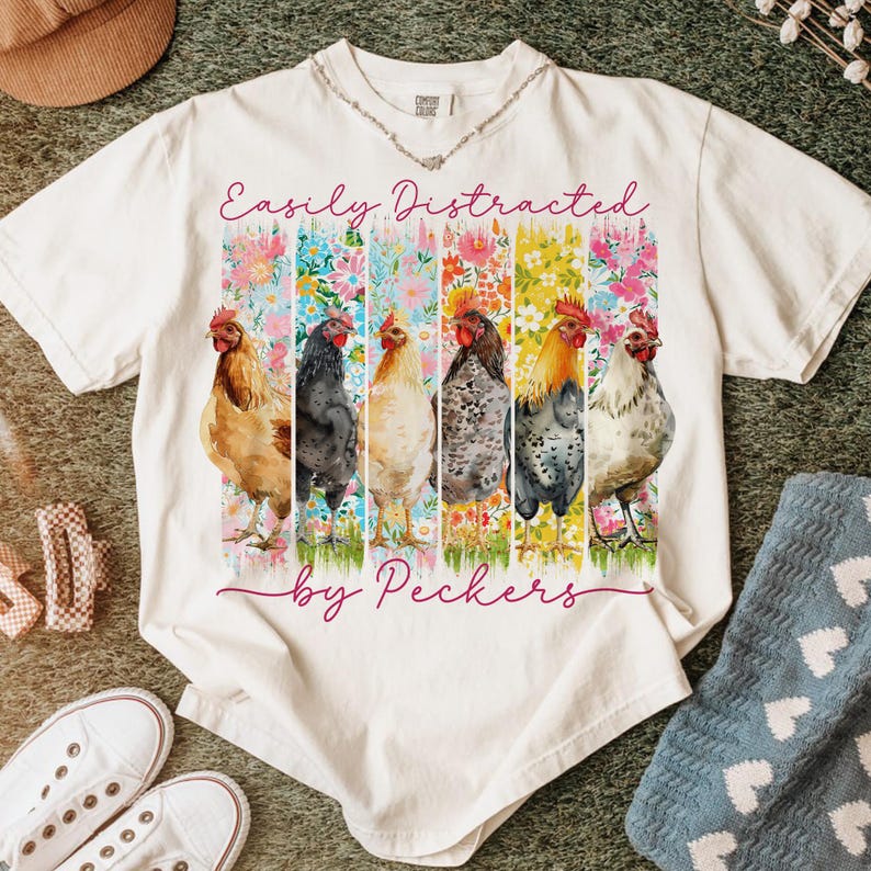 May include: White t-shirt with the text "Easily Distracted by Peckers." The shirt features a colorful graphic of six chickens in front of floral backgrounds. The chickens are in various colors, including brown, black, and white.