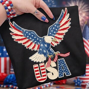 May include: Black tote bag featuring an embroidered eagle with outstretched wings, incorporating red, white, and blue elements. The word "USA" is prominently displayed below the eagle, using the same patriotic color scheme. The design is reminiscent of the American flag.