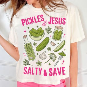 Peut inclure: T-shirt crème avec un motif graphique représentant des cornichons, un bocal de cornichons et d'autres illustrations liées aux cornichons. Le texte "PICKLES & JESUS SALTY & SAVE" est imprimé en rose.