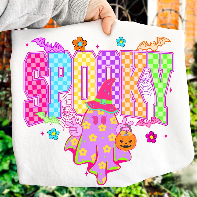 May include: White Halloween-themed tote bag with the word "SPOOKY" in colorful checkered letters. A cartoon ghost with a witch hat and a jack-o'-lantern bucket is featured. Bats and flowers decorate the design.