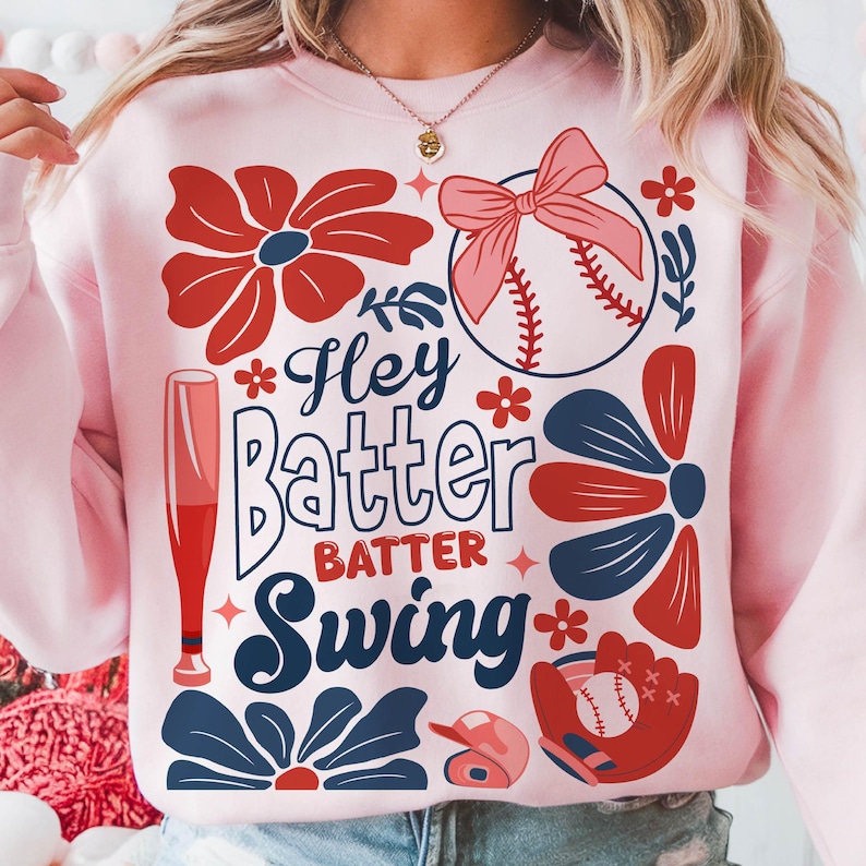 May include: Pink sweatshirt with a baseball theme. It features a baseball, bat, glove, and flowers. The text on the sweatshirt says "Hey Batter Batter Swing".