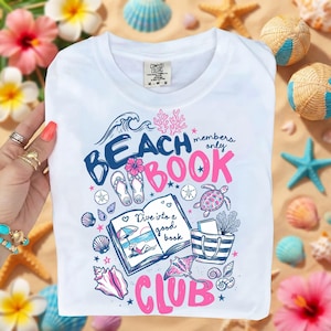May include: White t-shirt with a graphic design in blue and pink, featuring the words "Beach Book Club". The design includes illustrations of a book, seashells, a turtle, and other beach-themed elements. The shirt is surrounded by seashells and flowers.