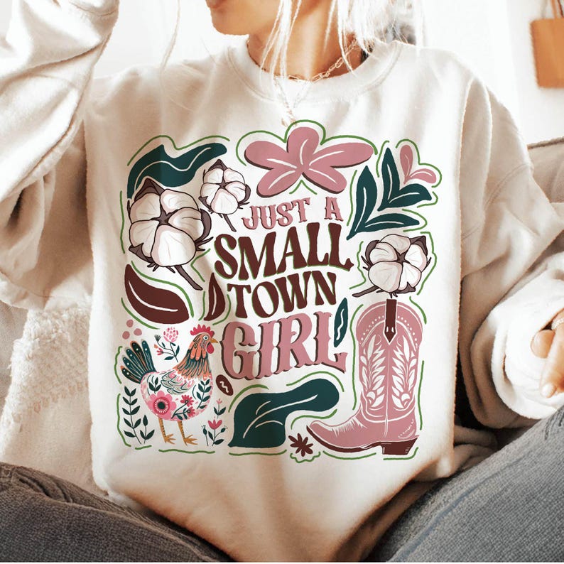 Pu&ograve; includere: Felpa color crema con un design grafico. Il design include il testo "Just a Small Town Girl", uno stivale da cowboy rosa, batuffoli di cotone, un gallo ed elementi floreali in rosa, verde e marrone.