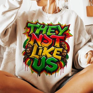 Może przedstawiać: Kremowa bluza z żywym wzorem graficznym. Projekt zawiera tekst "THEY NOT LIKE US" w zielonych, czerwonych i ż&oacute;łtych literach w stylu graffiti, a także ilustracje os&oacute;b z dredami.