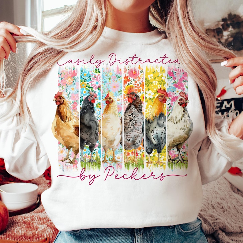 May include: White sweatshirt with a colorful graphic featuring six chickens in a row, each with a different color scheme. The graphic is set against floral backgrounds. Text reads "Easily Distracted by Peckers" in a cursive font.