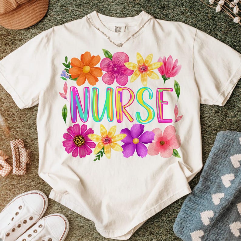 以下が含まれることがあります： クリーム色のTシャツ。カラフルな文字で「NURSE」の文字と花の模様がプリントされています。花はオレンジ、ピンク、黄色、紫の色合いです。柔らかい素材でできており、普段使いに適しています。