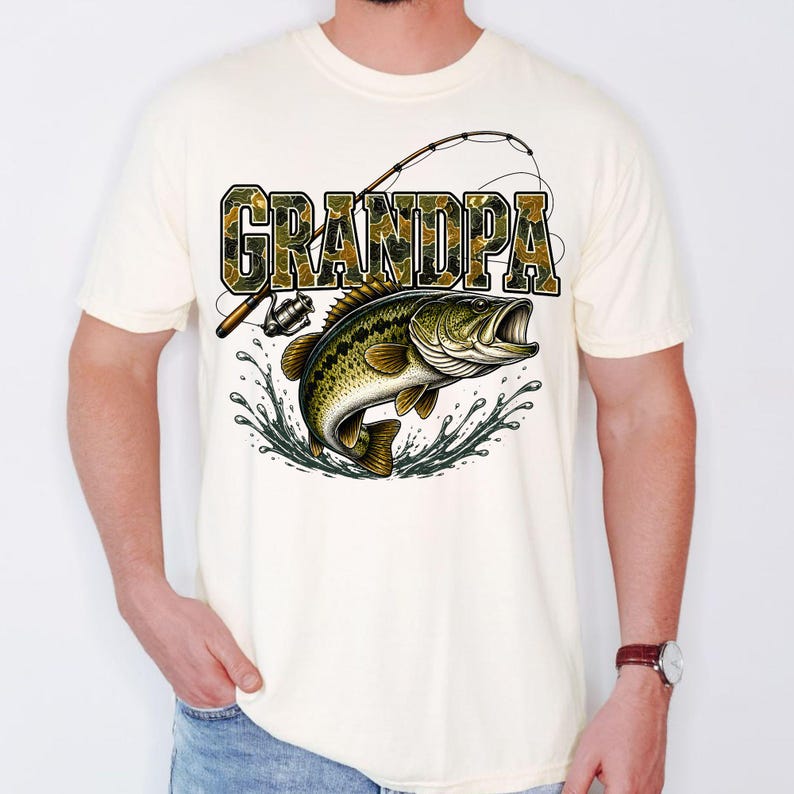K&ouml;nnte beinhalten: Cremefarbenes T-Shirt mit dem Wort "GRANDPA" in Tarnschrift &uuml;ber einer Grafik eines Gro&szlig;maulbasses, der aus dem Wasser springt. Eine Angelrute und -rolle sind ebenfalls in dem Design dargestellt.