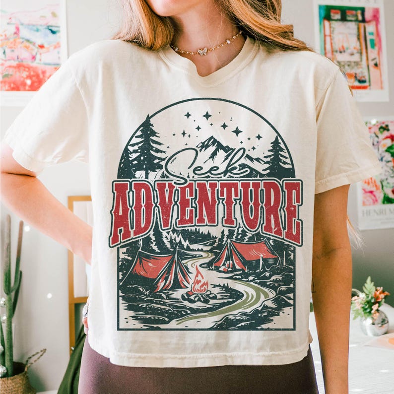 May include: A white t-shirt with a vintage-style graphic of a mountain scene with the text "Seek Adventure". The graphic features a campfire, two tents, and trees.
