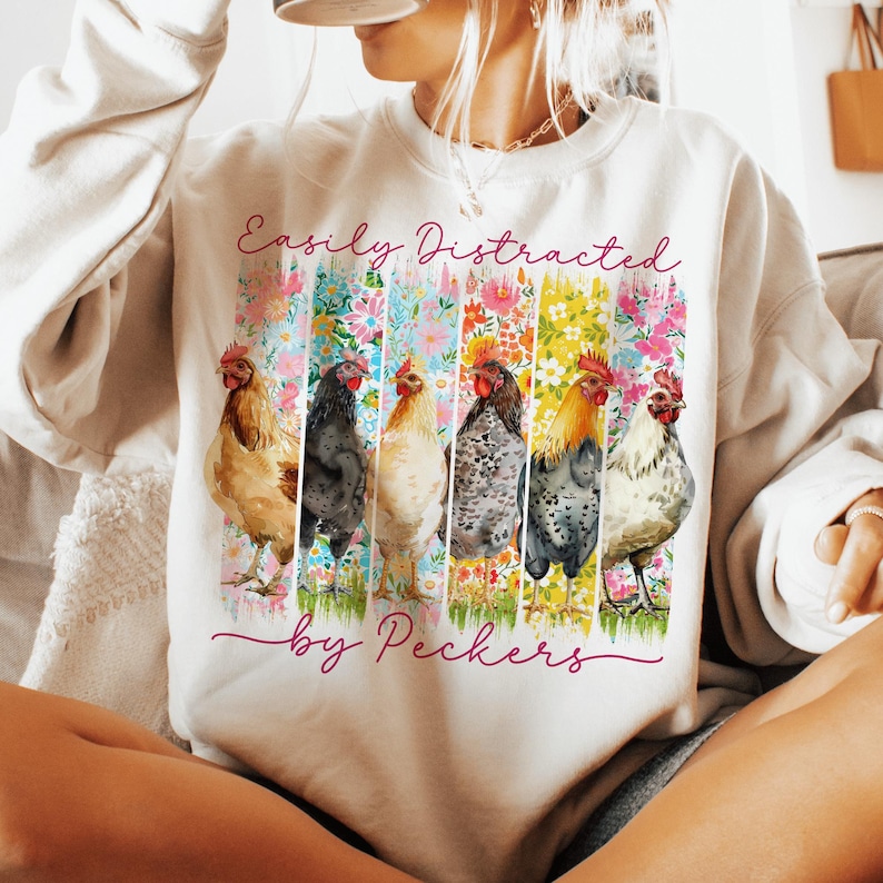 May include: Cream-colored sweatshirt with the text "Easily Distracted by Peckers" in pink script. The design features six watercolor chickens in front of colorful floral stripes. A casual, comfortable top.