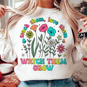 Puede incluir: Sudadera blanca con un gr&aacute;fico colorido de flores y el texto "Teach them, love them, watch them grow".