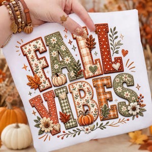 May include: A white t-shirt featuring the embroidered text FALL VIBES in large, decorative block letters. The letters are filled with various autumn-themed patterns including floral prints, plaid, and polka dots, accented with pumpkins and leaves.
