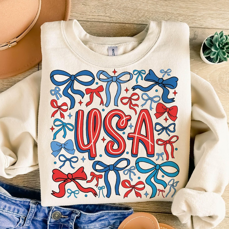 May include: Cream-colored sweatshirt with a patriotic design. The word "USA" is in red and blue, surrounded by red and blue bows. The design is centered on the front of the sweatshirt.