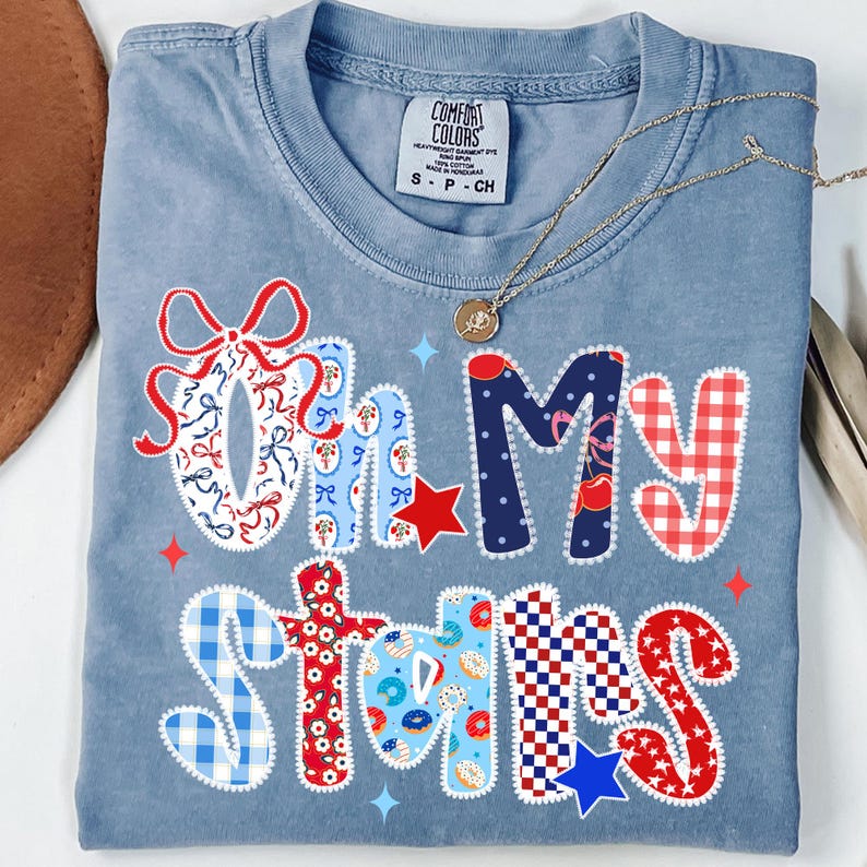 May include: A light blue t-shirt with the phrase "Oh My Stars" in a patchwork design. The letters are decorated with patterns including bows, stars, and gingham. A gold necklace with a pendant is draped across the shirt.