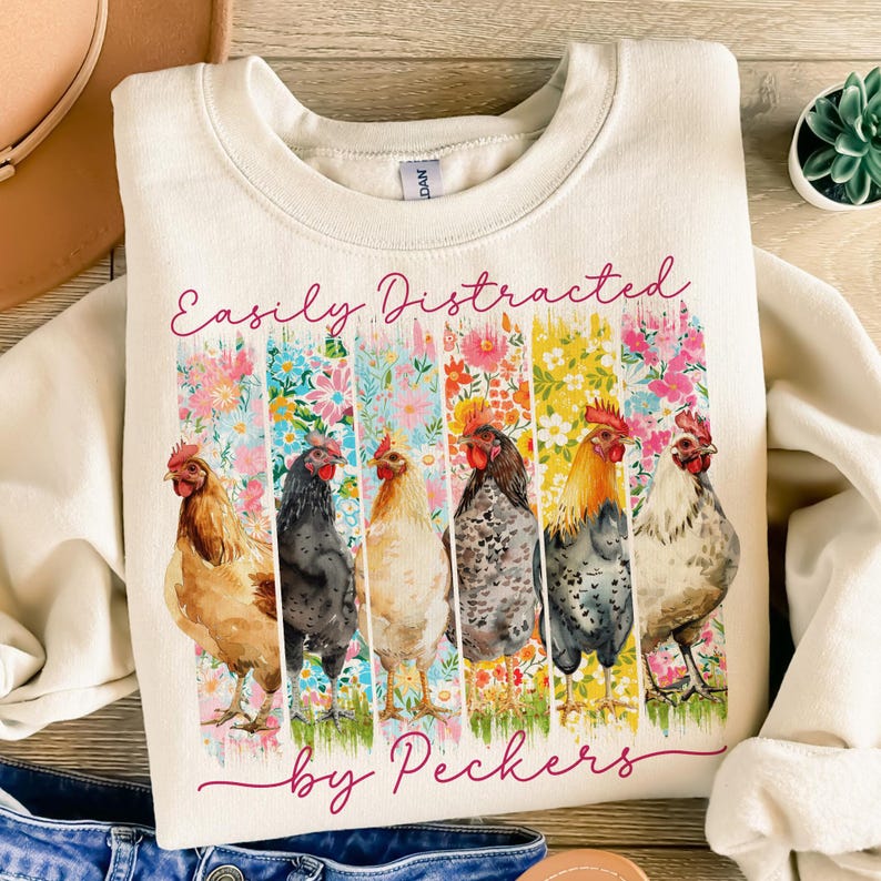 May include: Cream-colored sweatshirt with the text "Easily Distracted by Peckers." The design features six colorful chickens in front of vertical floral stripes. The sweatshirt is made of a soft material.