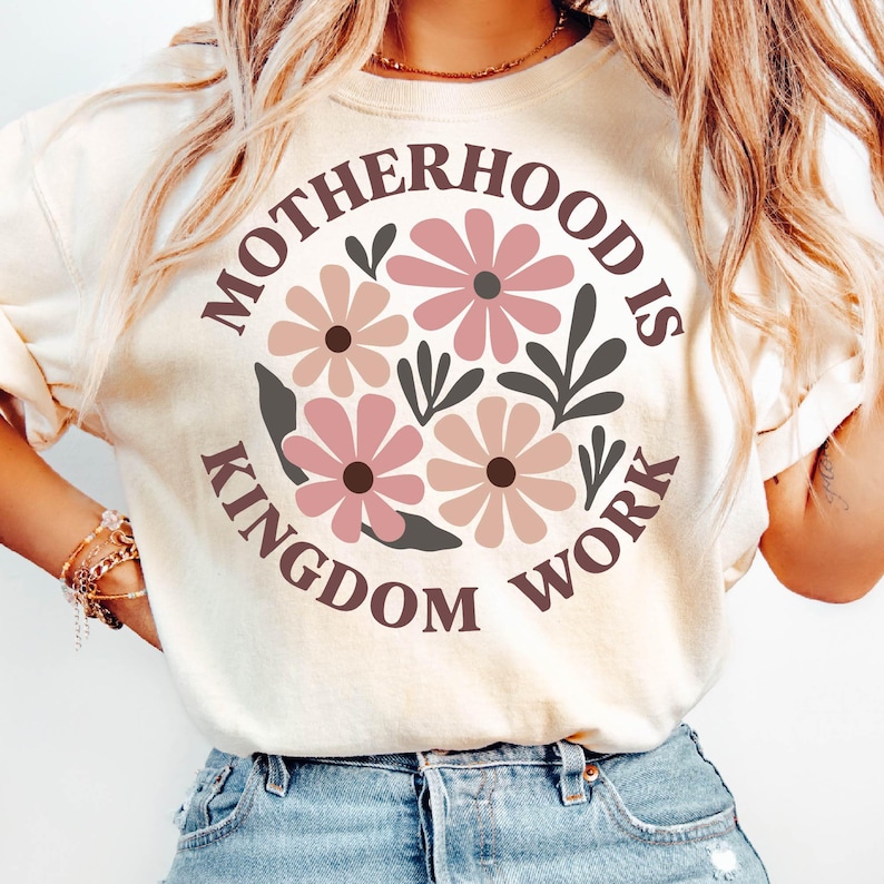 Puede incluir: Una camiseta blanca con un dise&ntilde;o floral rosa y el texto "Motherhood is Kingdom Work" en un c&iacute;rculo marr&oacute;n.