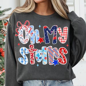 May include: Dark gray sweatshirt with the phrase "Oh My Stars" in a patchwork design. The letters feature patterns like gingham, stars, and bows in red, white, and blue. The sweatshirt has a crew neck and long sleeves.