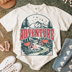 May include: White t-shirt with a vintage-style graphic of a campfire scene in the woods. The graphic includes a mountain range, trees, two tents, and a campfire. The text "Seek Adventure" is written in red above the graphic.
