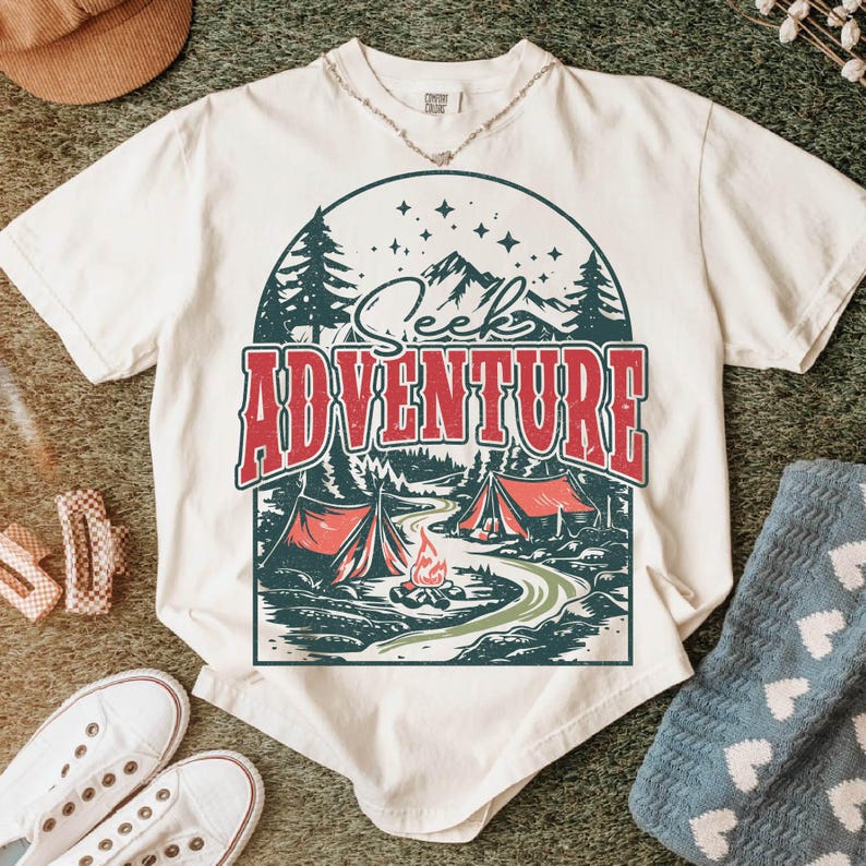 May include: White t-shirt with a vintage-style graphic of a campfire scene in the woods. The graphic includes a mountain range, trees, two tents, and a campfire. The text "Seek Adventure" is written in red above the graphic.