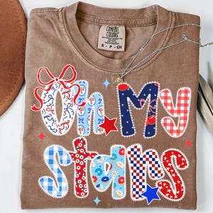 May include: A brown Comfort Colors t-shirt with the phrase "Oh My Stars" in a patchwork design. The letters are decorated with red, white, and blue patterns, including bows, stars, and gingham. A silver necklace rests on the shirt.