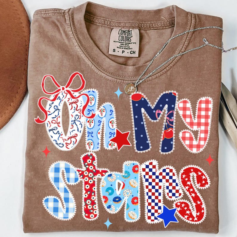 May include: A brown Comfort Colors t-shirt with the phrase "Oh My Stars" in a patchwork design. The letters are decorated with red, white, and blue patterns, including bows, stars, and gingham. A silver necklace rests on the shirt.