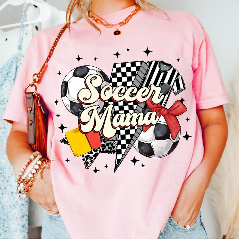 K&ouml;nnte beinhalten: Pinkes T-Shirt mit einem Grafikdesign aus einem Fu&szlig;ball, einem Schiedsrichter und einer karierten Flagge. Der Text "Soccer Mama" ist in einer Retro-Schrift.
