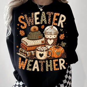 May include: A black sweatshirt featuring a graphic print with the text "SWEATER WEATHER". The design includes a stack of folded sweaters, a beanie with a pom-pom, a plastic cup with a lid, a mug of hot cocoa with marshmallows, and a pumpkin.