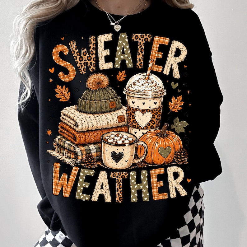 May include: A black sweatshirt featuring a graphic print with the text "SWEATER WEATHER". The design includes a stack of folded sweaters, a beanie with a pom-pom, a plastic cup with a lid, a mug of hot cocoa with marshmallows, and a pumpkin.