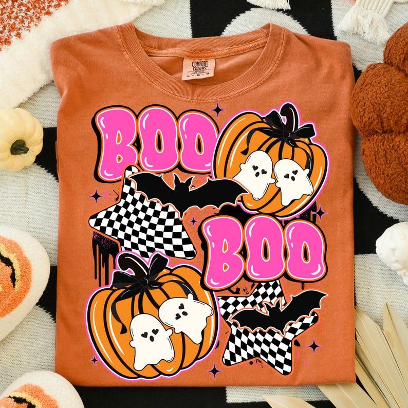 Puede incluir: Camiseta naranja Comfort Colors con un dise&ntilde;o de Halloween. El dise&ntilde;o incluye la palabra "BOO" en letras rosas, calabazas, fantasmas, murci&eacute;lagos y estrellas a cuadros. La camiseta es de cuello redondo.
