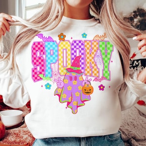 May include: White sweatshirt with a colorful "SPOOKY" graphic. The design features a ghost wearing a witch hat and holding a jack-o'-lantern, surrounded by bats, flowers, and spiderwebs. A fun, festive Halloween-themed top.