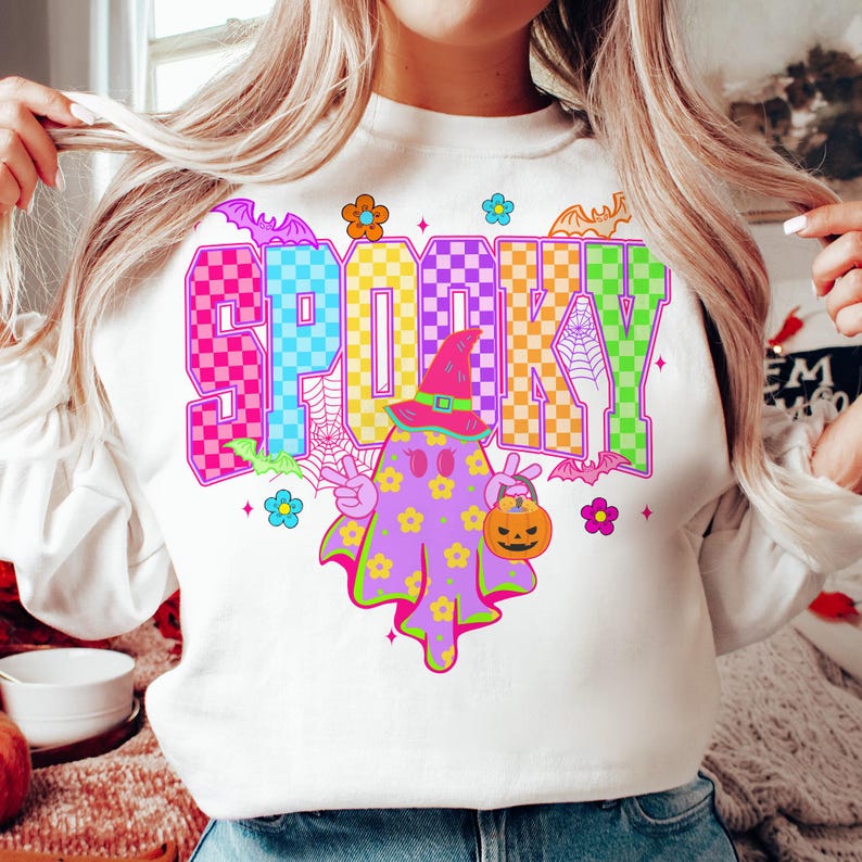 May include: White sweatshirt with a colorful "SPOOKY" graphic. The design features a ghost wearing a witch hat and holding a jack-o'-lantern, surrounded by bats, flowers, and spiderwebs. A fun, festive Halloween-themed top.