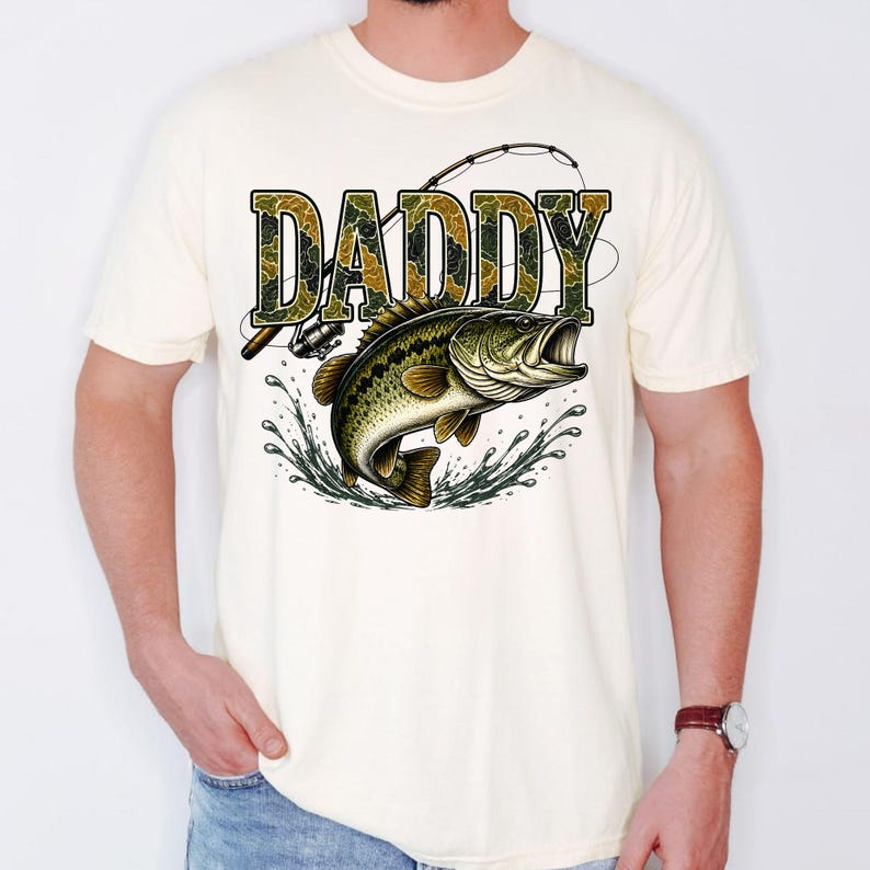 K&ouml;nnte beinhalten: Cremefarbenes T-Shirt mit einer Grafik eines Gro&szlig;maulbasses, der aus dem Wasser springt. Das Wort "DADDY" ist in einem Camouflage-Muster &uuml;ber dem Fisch aufgedruckt. Eine Angelrute und -rolle sind ebenfalls abgebildet.