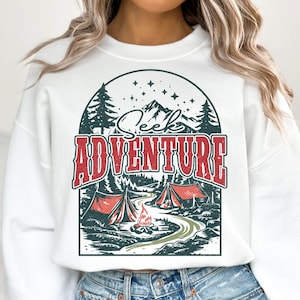 May include: White sweatshirt with a graphic print of a mountain scene with the text "Seek Adventure". The graphic features a campfire, two tents, and trees.
