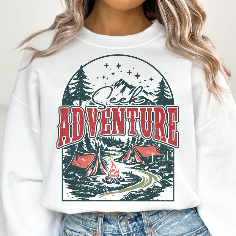 May include: White sweatshirt with a graphic print of a mountain scene with the text "Seek Adventure". The graphic features a campfire, two tents, and trees.