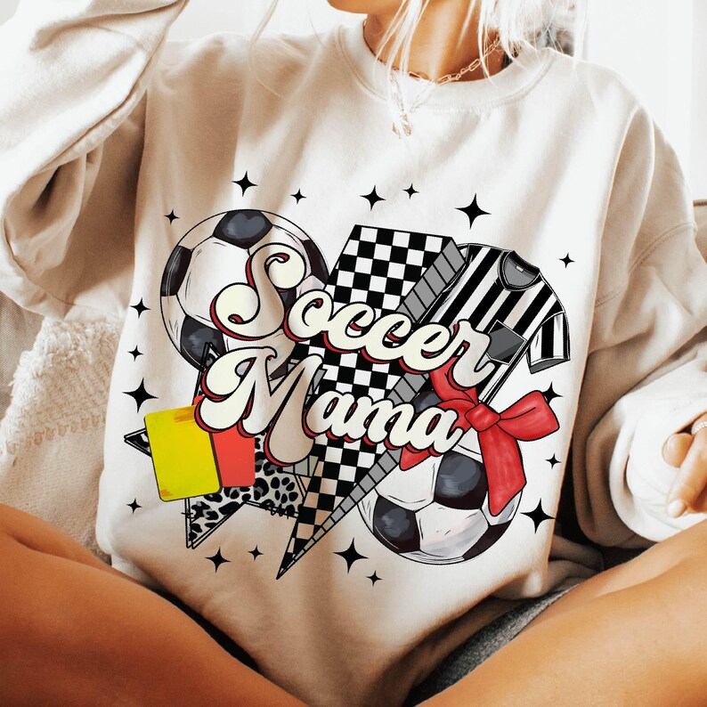 K&ouml;nnte beinhalten: Wei&szlig;er Pullover mit einem Grafikdesign aus einem Fu&szlig;ball, einer gelben Karte, einer karierten Flagge und einem Fu&szlig;balltrikot. Die Grafik ist herzf&ouml;rmig und sagt "Soccer Mama" in einer Retro-Schrift.